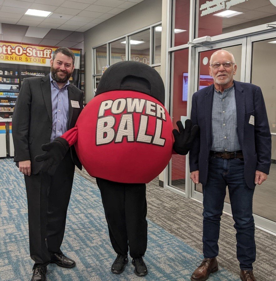 Picture2_Two-men-of-average-height-standing-next-to-a-red-power-ball-mascot-wearking-a-black-baseball-cap-turned-backwards.-They-are-in-front-of-a-lottery-store-facade